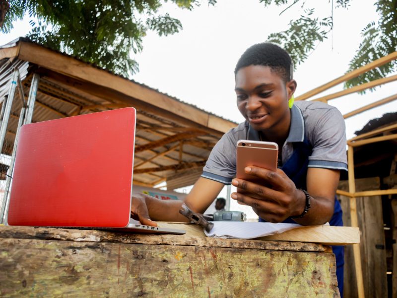 african carpenter holding a phone and pen wearing an apron working with his phone and laptop